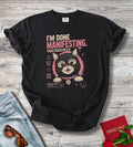 A black graphic t-shirt laid flat on a gray wood background, styled with light blue jeans, sunglasses, a red book, and red roses. The t-shirt features a graphic of a cartoon cat inside a pink circular frame with the text 'I'M DONE MANIFESTING.