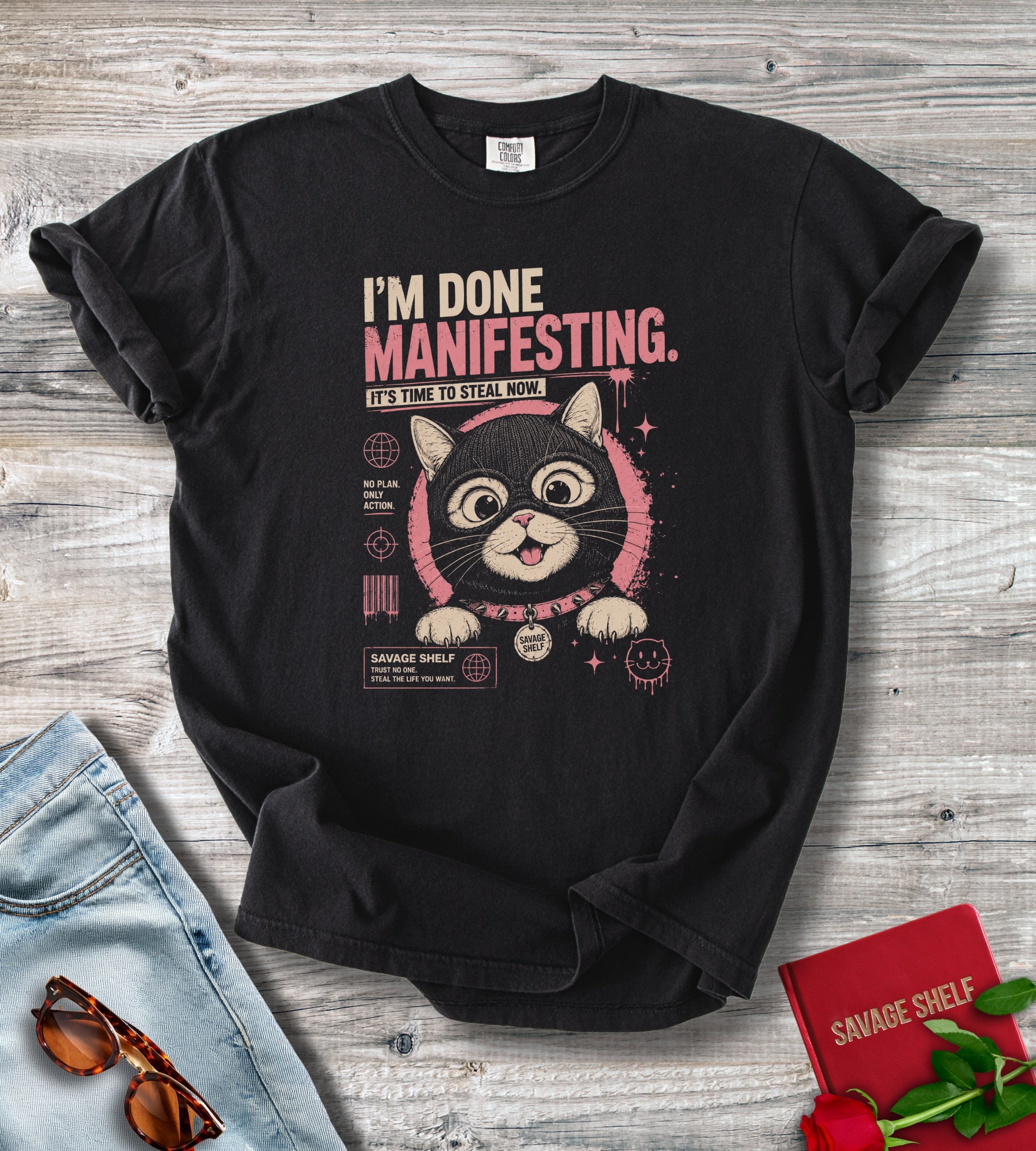 A black graphic t-shirt laid flat on a gray wood background, styled with light blue jeans, sunglasses, a red book, and red roses. The t-shirt features a graphic of a cartoon cat inside a pink circular frame with the text 'I'M DONE MANIFESTING.