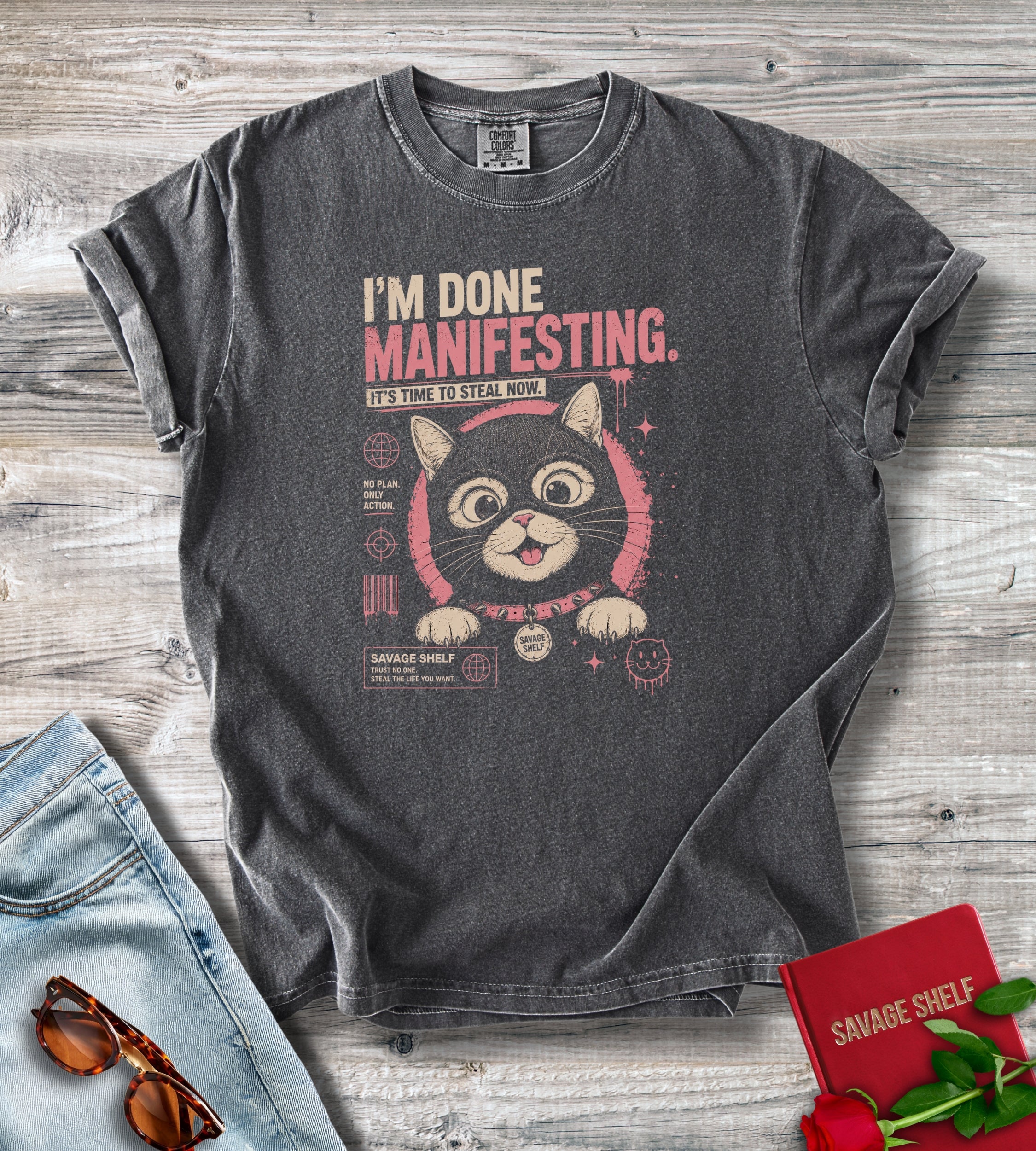 A dark gray graphic t-shirt laid flat on a rustic wood background, styled with light blue jeans, sunglasses, and a red notebook. The t-shirt features a distressed graphic of a cat with the text 'I'M DONE MANIFESTING.