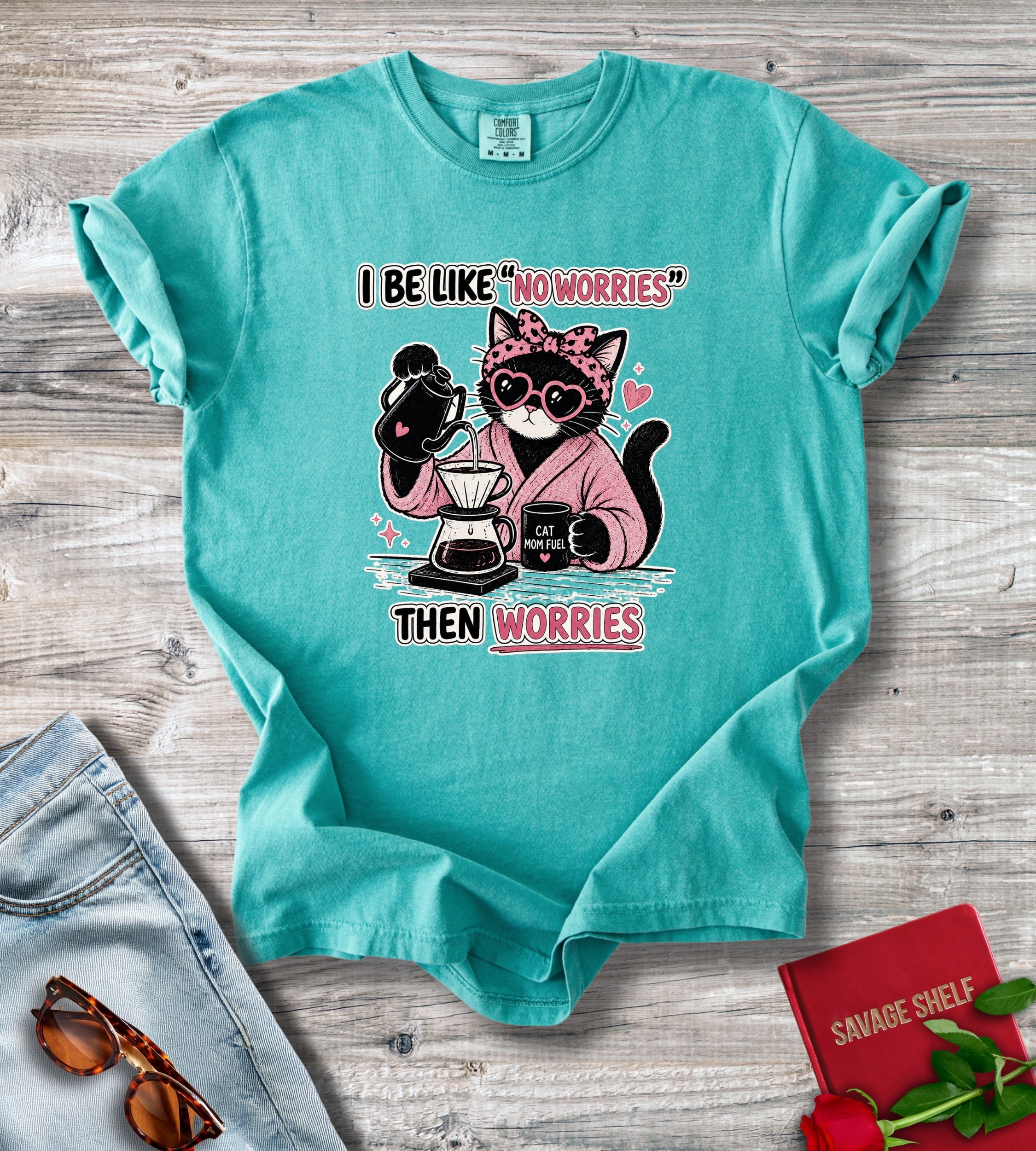 A turquoise graphic t-shirt laid flat on a gray wood background, styled with a pair of blue jeans, sunglasses, and a red book. The t-shirt features a cartoon cat wearing sunglasses and a pink hoodie, holding coffee equipment, with the text 'I BE LIKE "NO WORRIES" THEN WORRIES'.