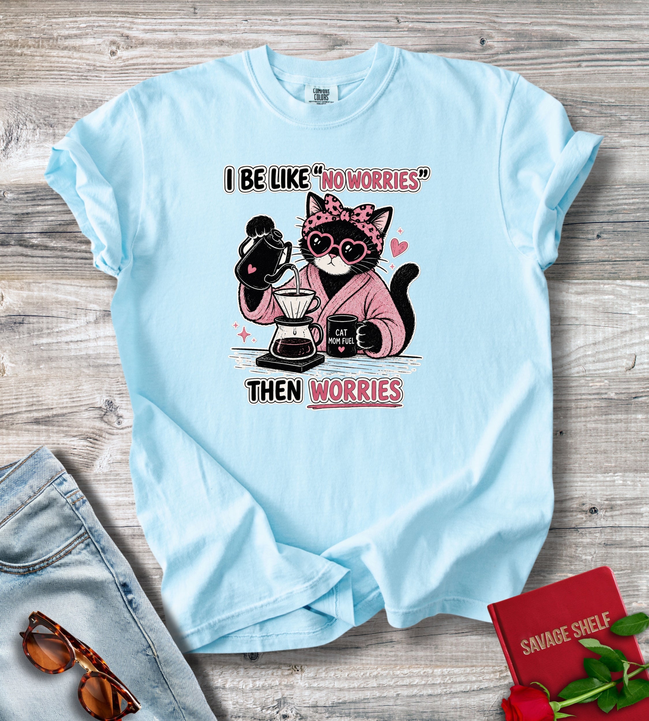 A light blue graphic t-shirt laid flat on a gray wood background, styled with sunglasses, denim shorts, and a red book. The t-shirt features a cartoon illustration of a pink cat wearing sunglasses and a polka-dot bow headband, holding coffee makers and a pour-over coffee dripper, with the text 'I BE LIKE NO WORRIES THEN WORRIES'.