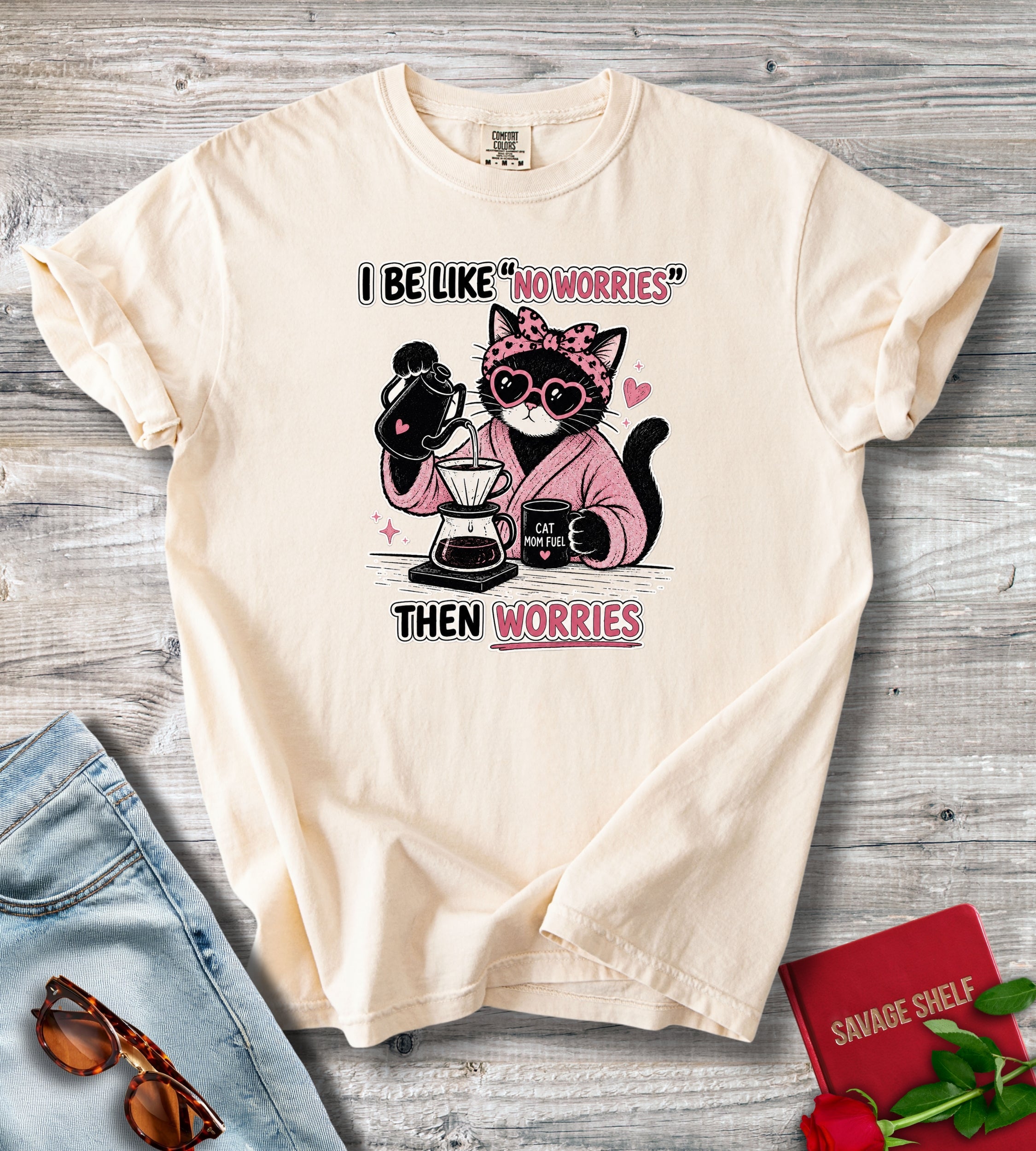 A cream-colored short-sleeve t-shirt with a graphic of a cat wearing sunglasses and a pink robe while making coffee, with the text 'I BE LIKE "NO WORRIES" THEN WORRIES'. The shirt is displayed in a flat-lay arrangement on a gray wood background, styled with blue jeans, sunglasses, a red notebook, and red roses.