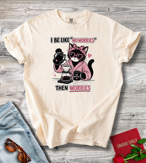 A cream-colored short-sleeve t-shirt with a graphic of a cat wearing sunglasses and a pink robe while making coffee, with the text 'I BE LIKE "NO WORRIES" THEN WORRIES'. The shirt is displayed in a flat-lay arrangement on a gray wood background, styled with blue jeans, sunglasses, a red notebook, and red roses.