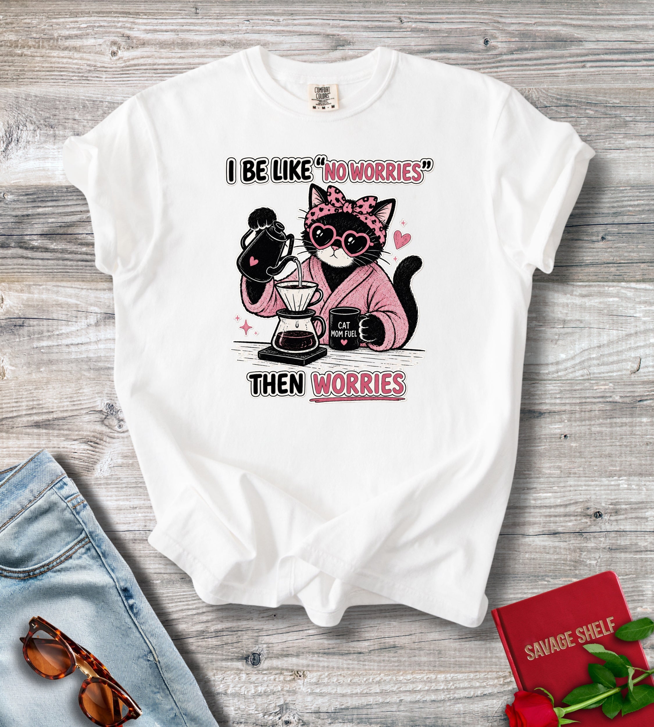 A white short-sleeve graphic T-shirt laid flat on a gray wood surface, styled with jeans, sunglasses, a red book, and a rose. The shirt has a cartoon cat wearing sunglasses and a pink robe while making coffee, with the text 'I BE LIKE "NO WORRIES" THEN WORRIES'.