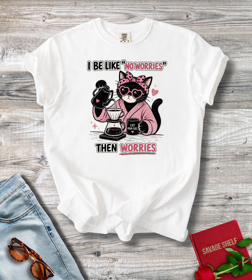 A white short-sleeve graphic T-shirt laid flat on a gray wood surface, styled with jeans, sunglasses, a red book, and a rose. The shirt has a cartoon cat wearing sunglasses and a pink robe while making coffee, with the text 'I BE LIKE "NO WORRIES" THEN WORRIES'.