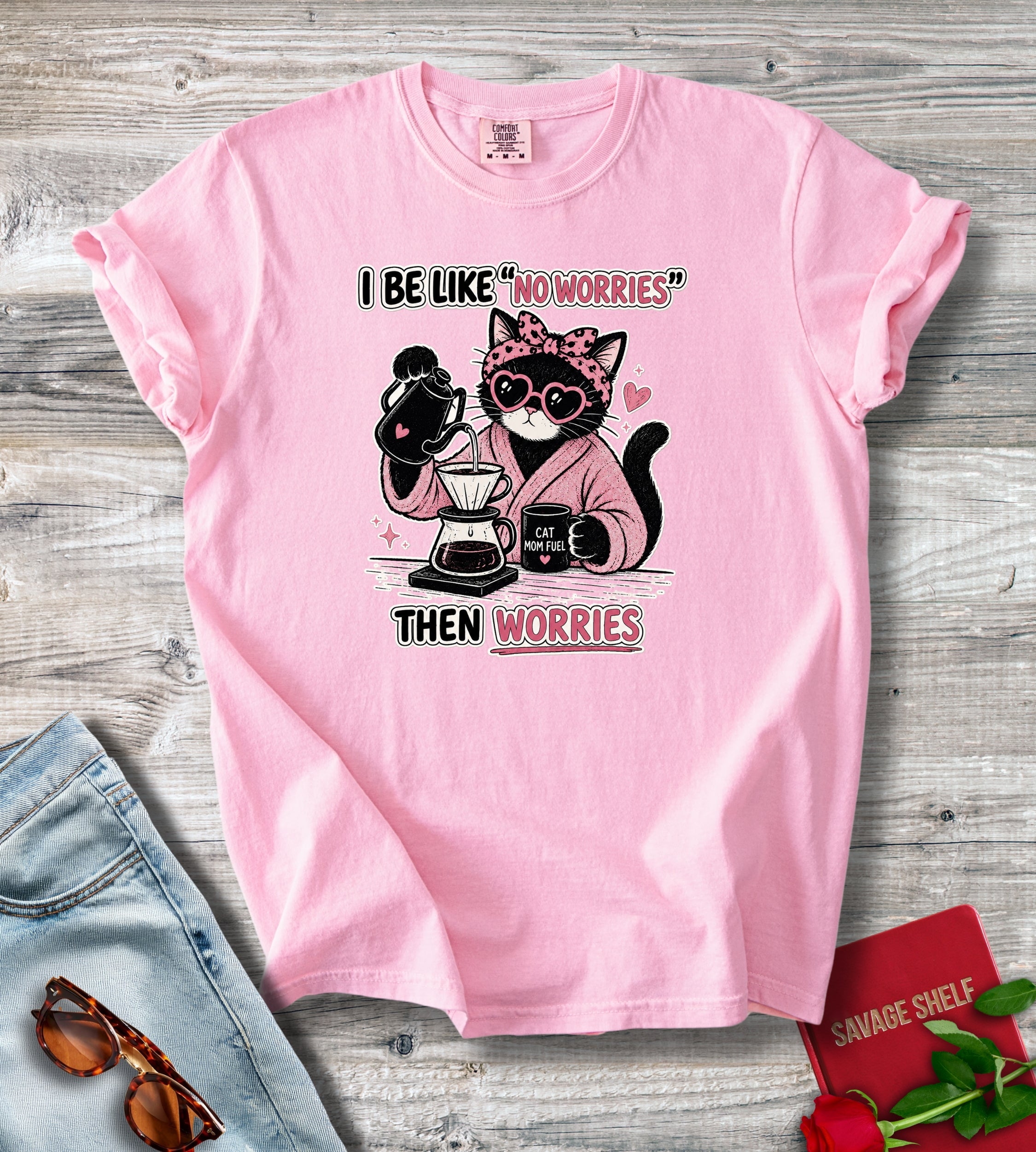 A pink short-sleeve graphic T-shirt laid flat on a rustic wood surface, with a cartoon cat wearing sunglasses and holding coffee cups, plus the text 'I BE LIKE NO WORRIES THEN WORRIES.' Jeans, sunglasses, and a red notebook are styled around the shirt.