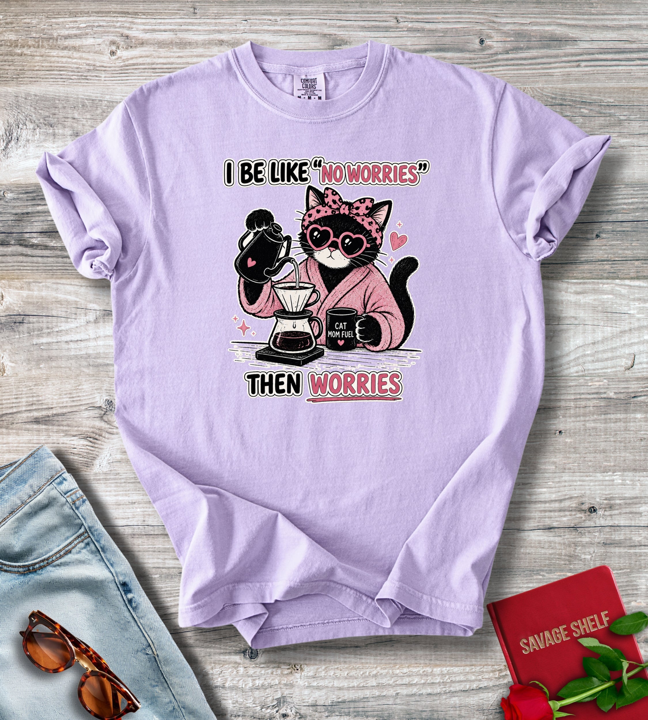 A lavender short-sleeve graphic T-shirt laid flat on a rustic wood surface, styled with jeans, sunglasses, a red book, and a rose. The shirt features a cartoon cat wearing sunglasses and a pink robe while pouring coffee, with the text 'I BE LIKE "NO WORRIES" THEN WORRIES'.