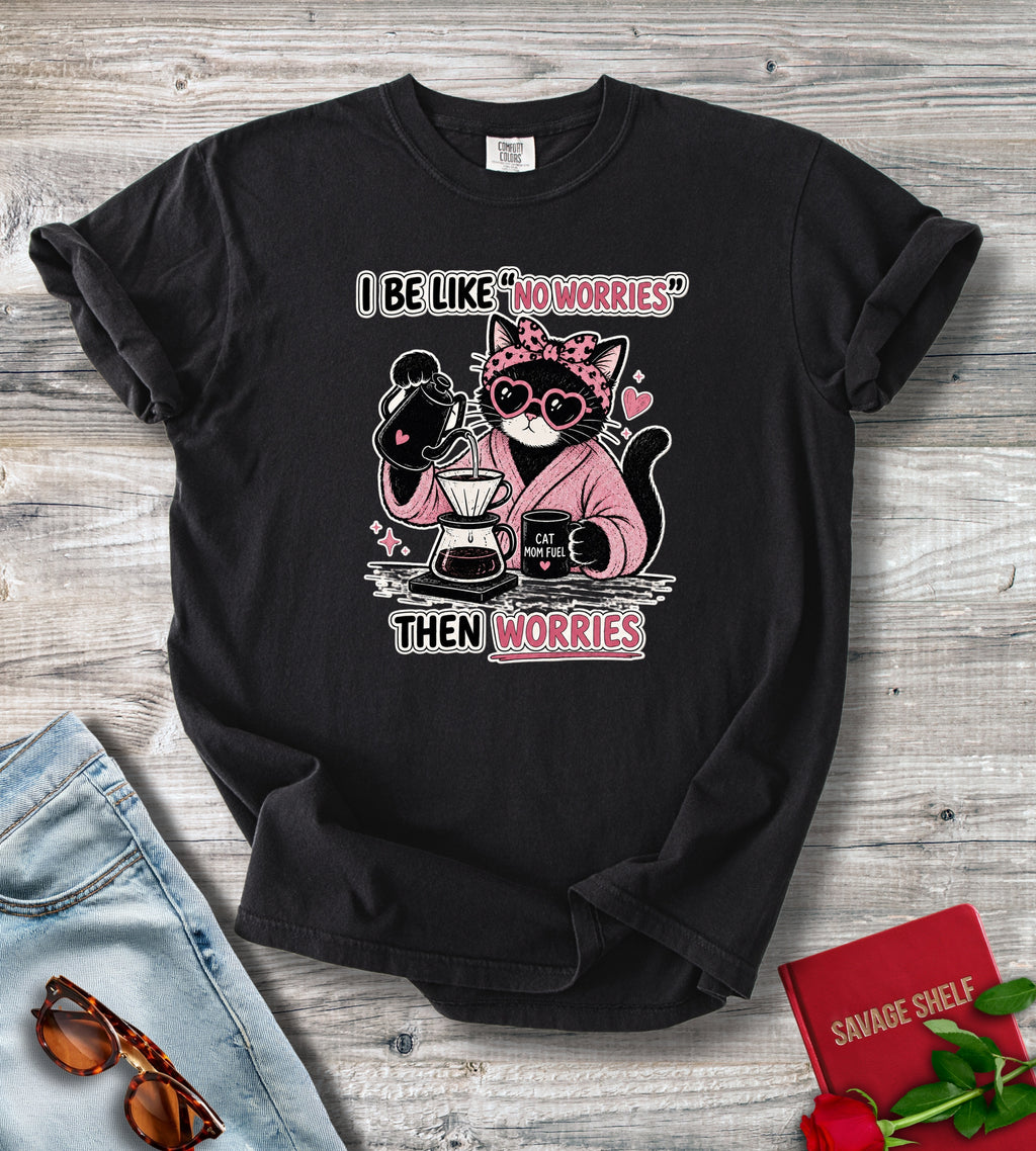 A black short-sleeve graphic T-shirt laid flat on a light wood surface, styled with jeans, sunglasses, a red book, and red roses. The shirt has a printed cartoon cat wearing heart-shaped sunglasses and a pink hoodie, holding coffee equipment, with the text 'I BE LIKE "NOWORRIES" THEN WORRIES'.