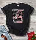 A black short-sleeve graphic T-shirt laid flat on a light wood surface, styled with jeans, sunglasses, a red book, and red roses. The shirt has a printed cartoon cat wearing heart-shaped sunglasses and a pink hoodie, holding coffee equipment, with the text 'I BE LIKE "NOWORRIES" THEN WORRIES'.