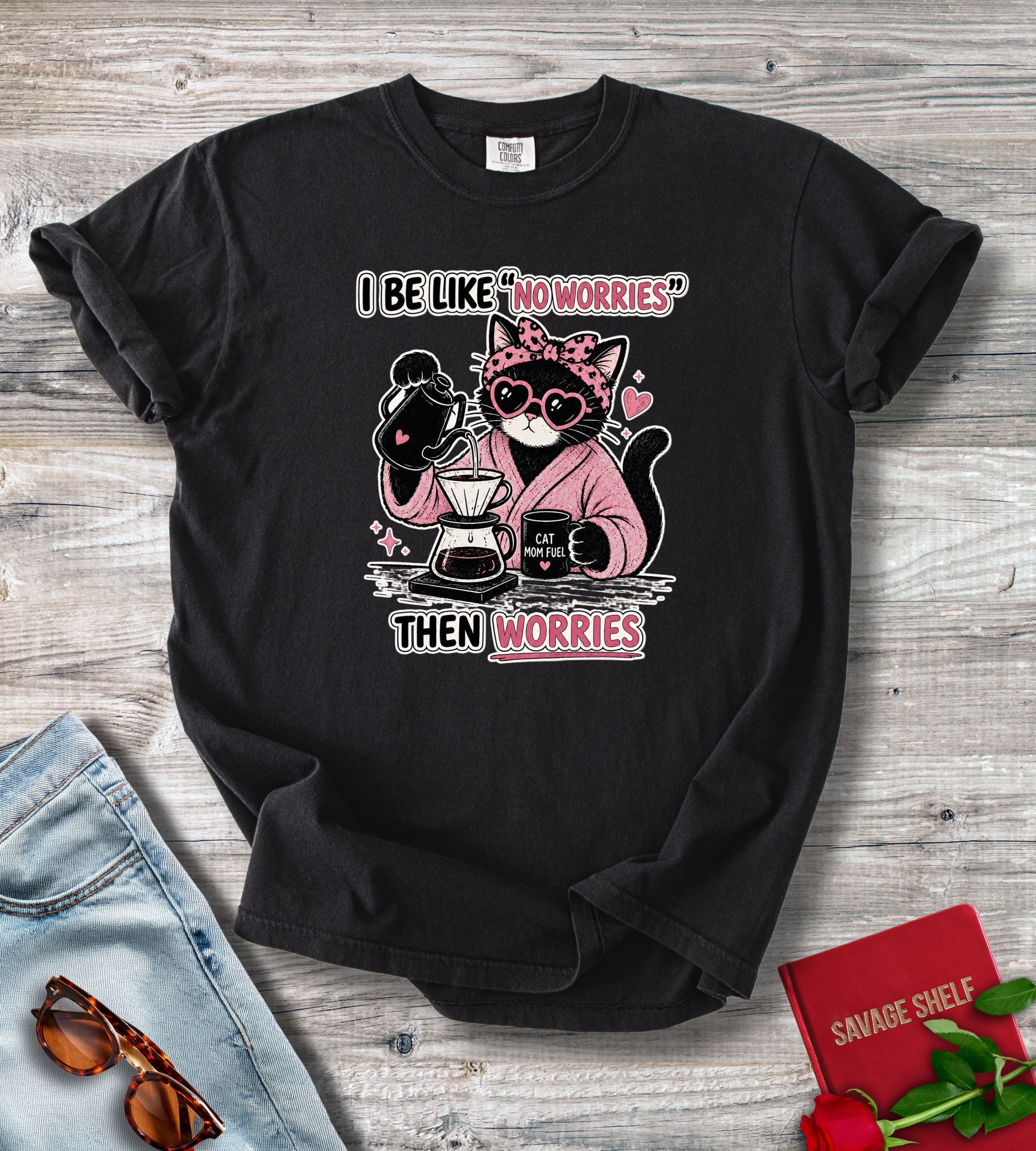 A black short-sleeve graphic T-shirt laid flat on a light wood surface, styled with jeans, sunglasses, a red book, and red roses. The shirt has a printed cartoon cat wearing heart-shaped sunglasses and a pink hoodie, holding coffee equipment, with the text 'I BE LIKE "NOWORRIES" THEN WORRIES'.