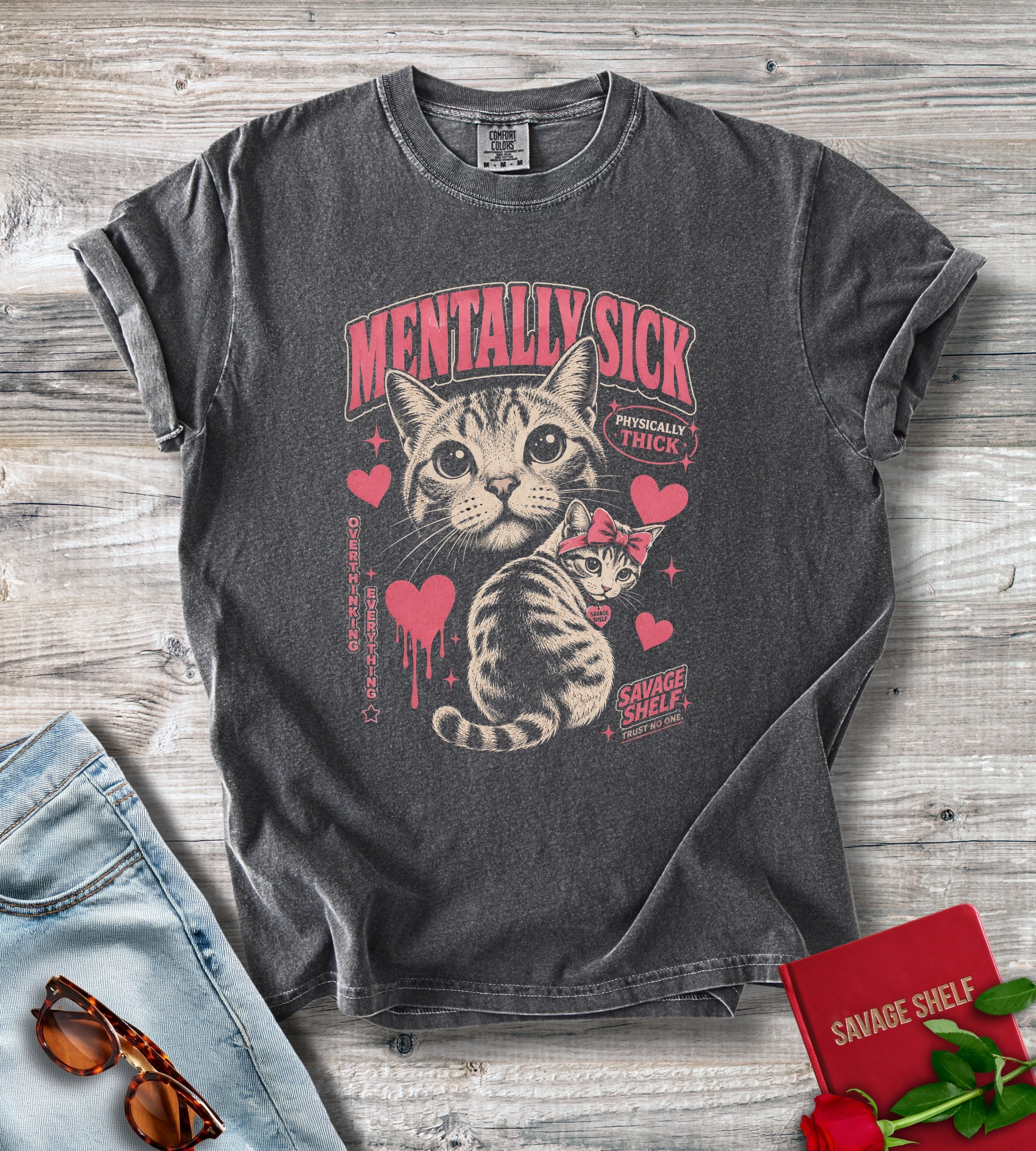 A dark gray graphic t-shirt laid flat on a rustic wood background, featuring a vintage-style illustration of a tabby cat with hearts and the text 'MENTALLY SICK' and 'SAVAGE SHELF'. Blue jeans, sunglasses, and a red notebook with a rose are styled around the shirt.