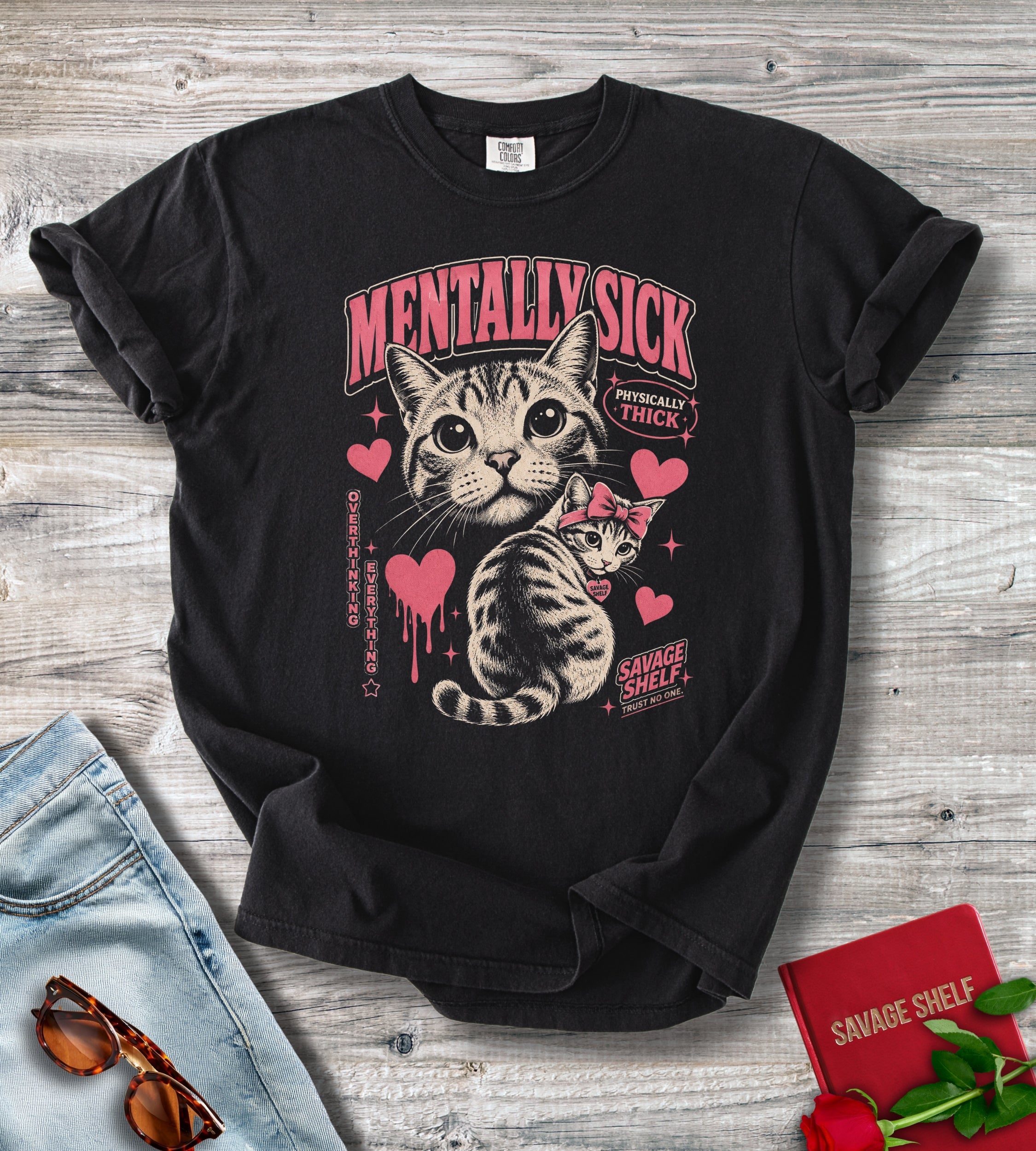 A black graphic t-shirt featuring a cat illustration and the text 'MENTALLY SICK' and 'SAVAGE SHELF' in pink and beige, styled in a flat-lay on a gray wood background with jeans, sunglasses, and a red book.