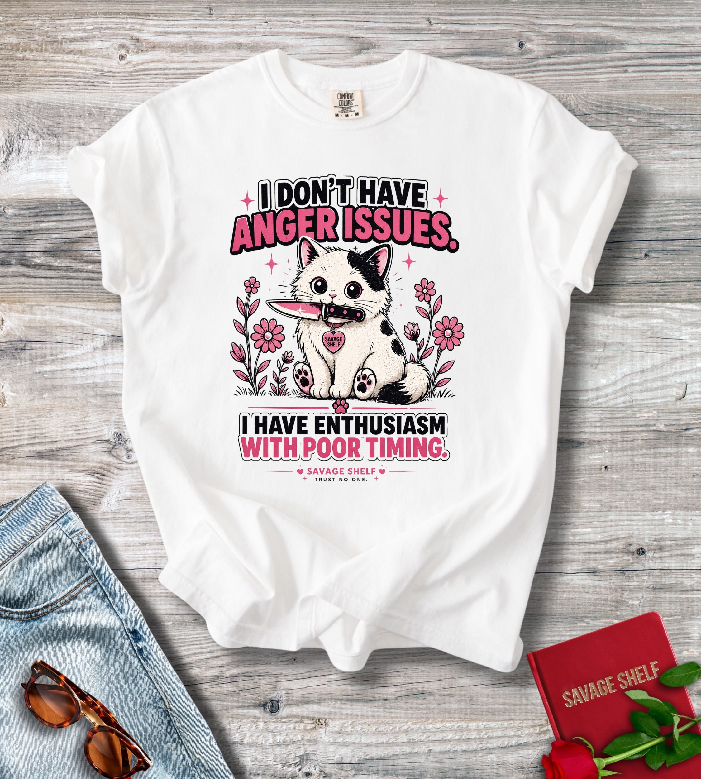 A white graphic t-shirt laid flat on a gray wood background, styled with blue jeans, sunglasses, a red book, and red roses. The t-shirt features a cartoon cat with a pink tongue sticking out, surrounded by pink flowers, with text that reads 'I DON'T HAVE ANGER ISSUES.