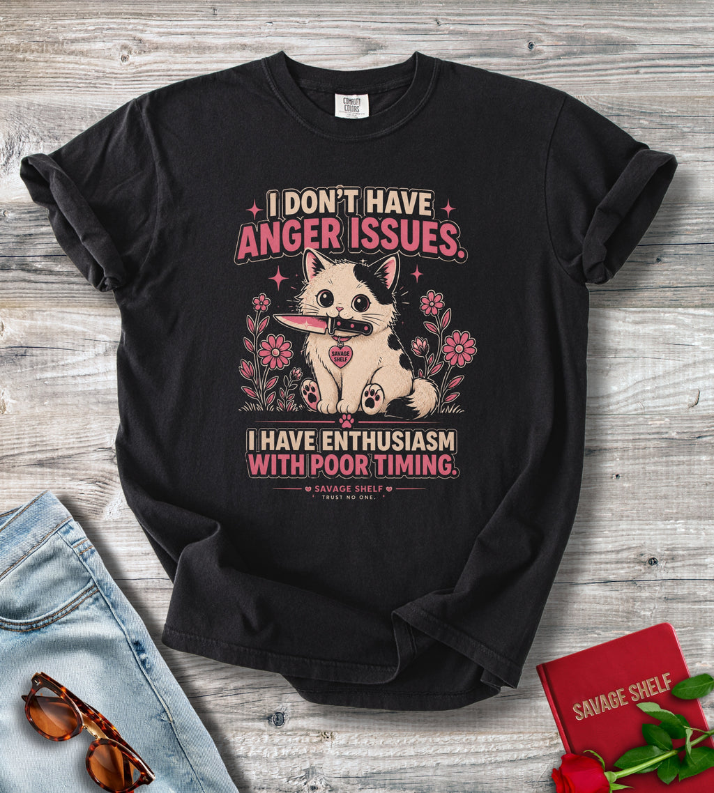 A black graphic t-shirt laid flat on a light gray wood background, styled with a pair of blue jeans, sunglasses, a red book, and a rose. The t-shirt features a cartoon cat holding a knife with the text 'I DON'T HAVE ANGER ISSUES.