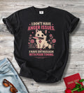 A black graphic t-shirt laid flat on a light gray wood background, styled with a pair of blue jeans, sunglasses, a red book, and a rose. The t-shirt features a cartoon cat holding a knife with the text 'I DON'T HAVE ANGER ISSUES.