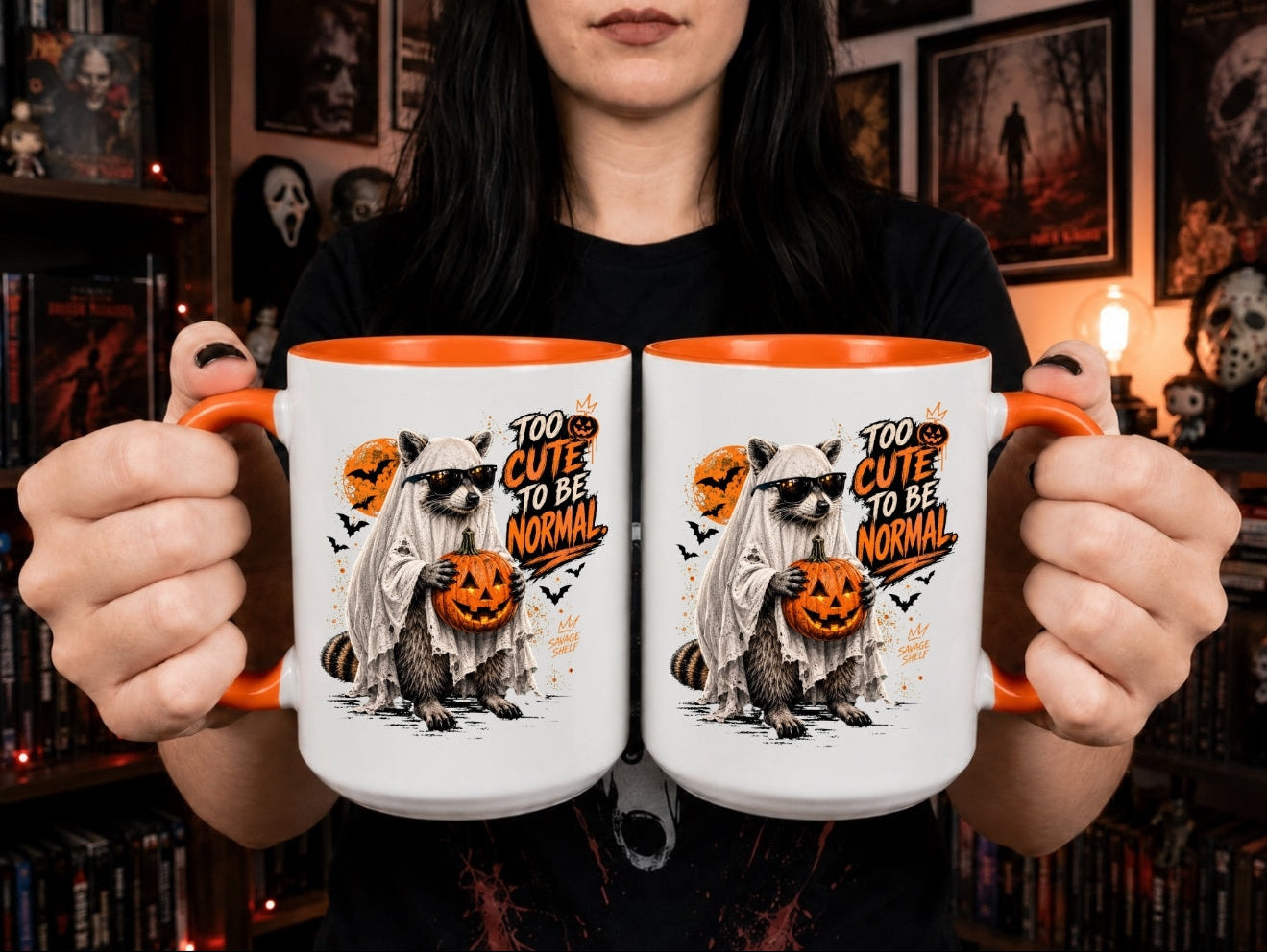 A person holds two white mugs with orange interiors and handles in front of a dark bookshelf. The mugs are printed with a Halloween-themed raccoon illustration in a ghost costume holding a jack-o'-lantern, with the text 'TOO CUTE TO BE NORMAL' and small bats.