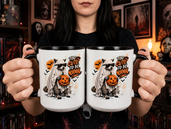 A person holds two white ceramic mugs with black handles and interiors. The mugs feature a Halloween graphic of a raccoon in a white costume holding a jack-o'-lantern, with the text 'TOO CUTE TO BE NORMAL' in orange and black.