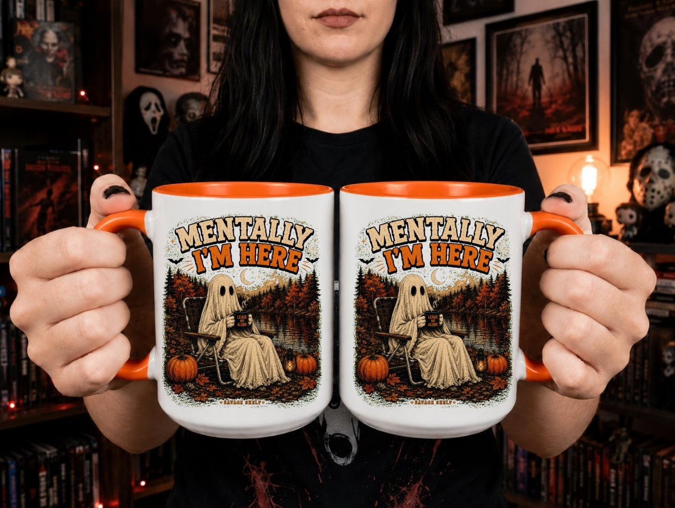A person holds two white ceramic mugs with orange interiors and handles, featuring a Halloween-themed graphic of a ghostly figure sitting on a pumpkin lawn with the text 'MENTALLY I'M HERE'. The background shows a cozy, dark bookshelf and framed wall art.