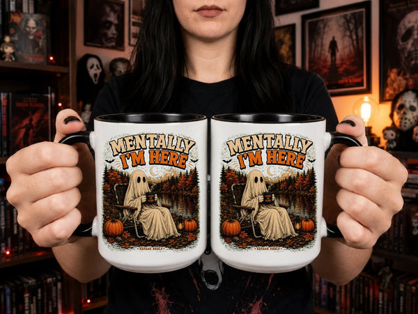A person holds two white ceramic mugs with black handles in front of a dark, spooky room background. The mugs feature a Halloween graphic of a ghost sitting in a chair by a lake with pumpkins, and the text 'MENTALLY I'M HERE' in a distressed font.