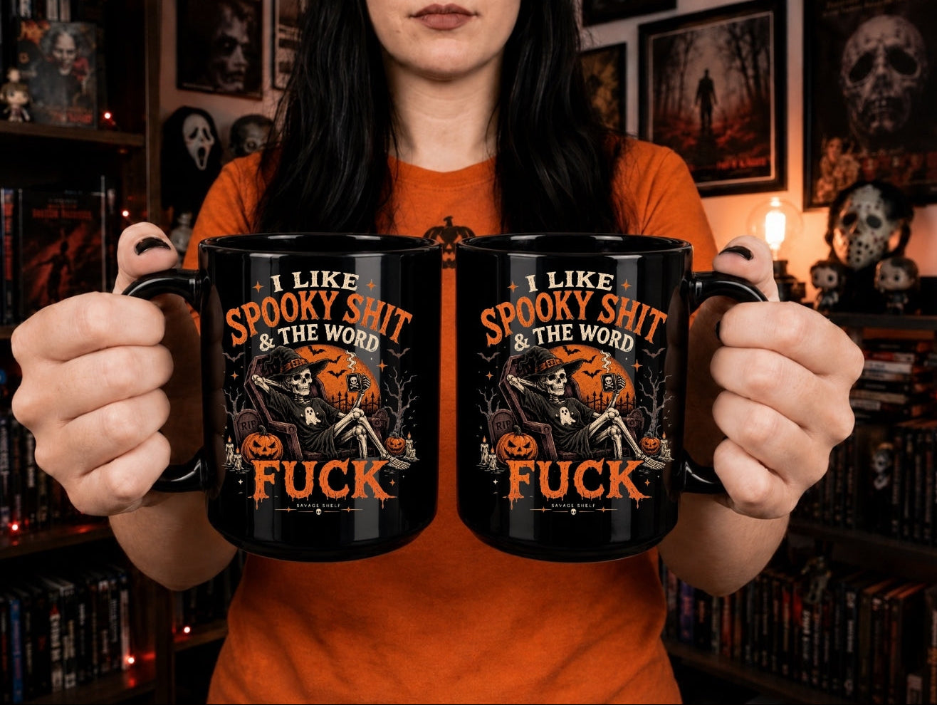 A person in an orange shirt holds two black mugs with a Halloween graphic that reads 'I LIKE SPOOKY SHIT & THE WORD FUCK' in orange text with a skeleton and pumpkin illustration, against a dark bookshelf background.