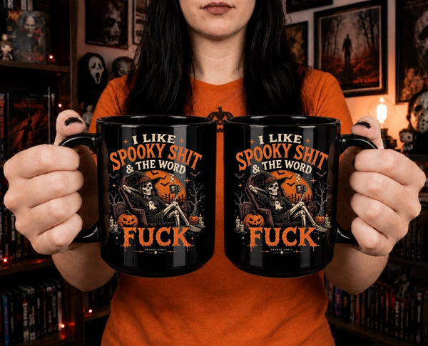 A person in an orange shirt holds two black mugs with a Halloween graphic that reads 'I LIKE SPOOKY SHIT & THE WORD FUCK' in orange text with a skeleton and pumpkin illustration, against a dark bookshelf background.