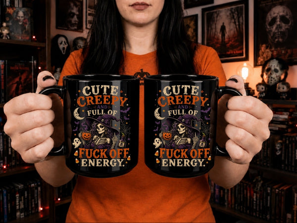 A person in an orange shirt holds up two black mugs with a Halloween-themed graphic that reads 'CUTE CREEPY AND FULL OF FUCK OFF ENERGY' with a skeleton, moon, and pumpkins on the mugs, set against a dark bookshelf background.