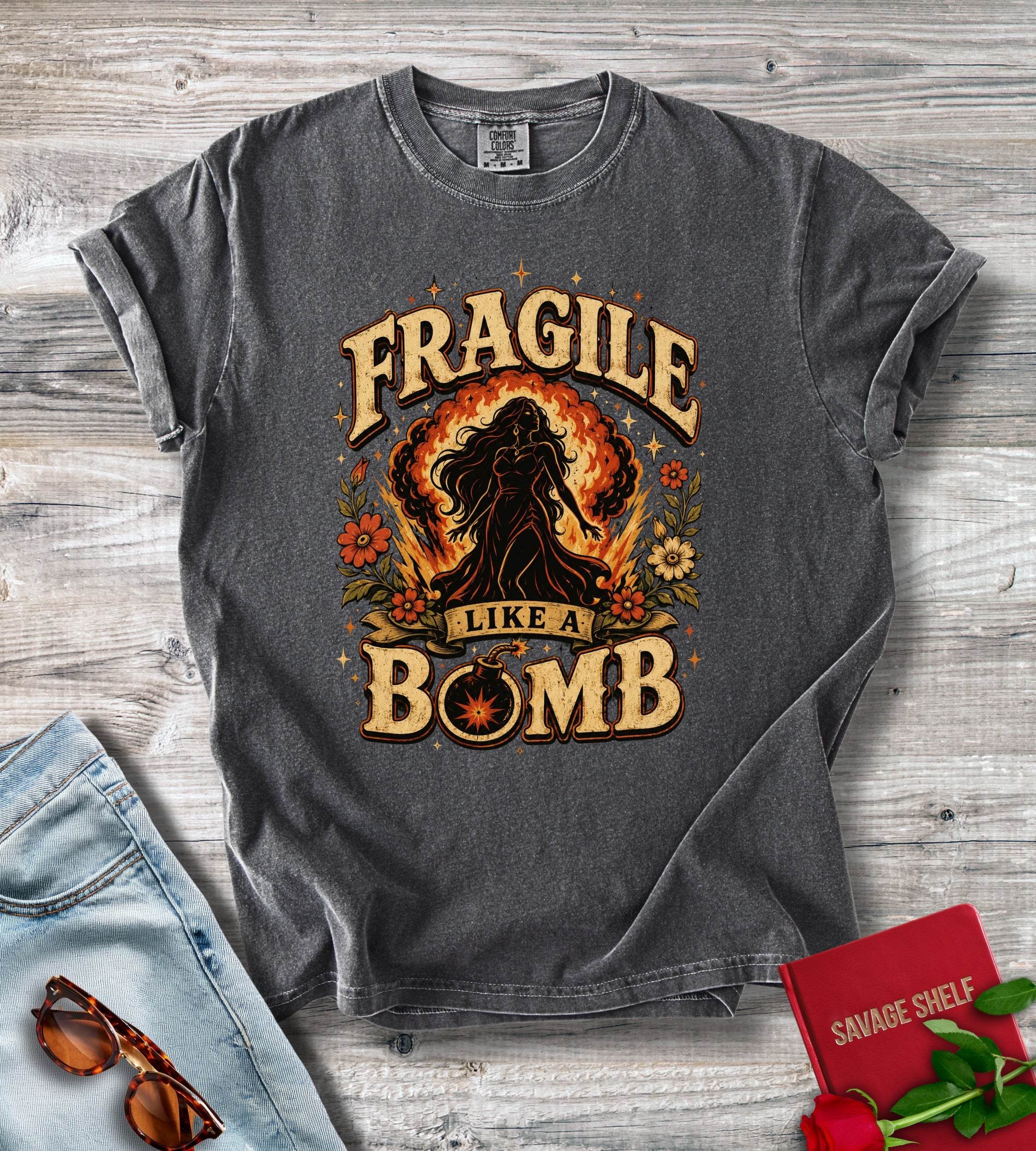 A dark gray graphic t-shirt laid flat on a light wood background, styled with light blue jeans, sunglasses, a red book, and red roses. The t-shirt features a graphic with the text 'FRAGILE LIKE A BOMB' and a silhouette of a woman surrounded by flames.