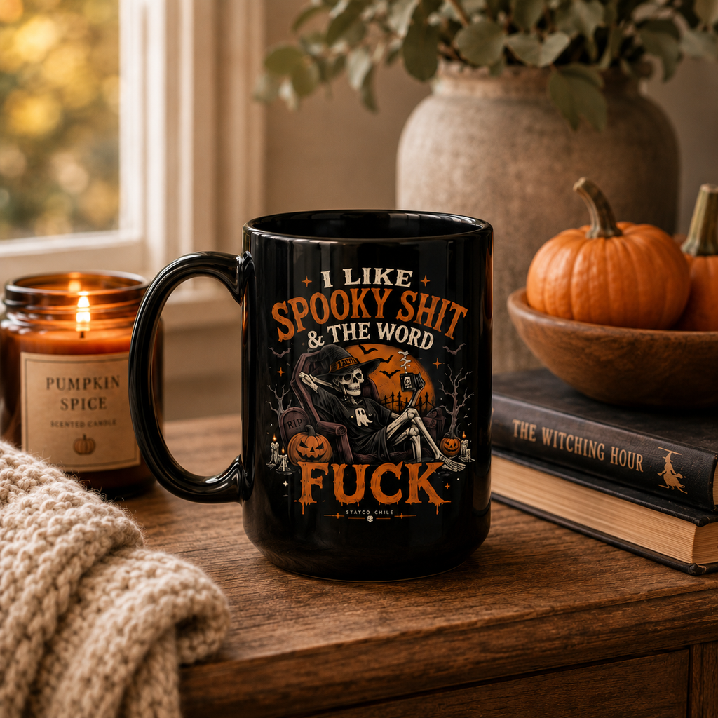 A black ceramic mug with a Halloween-themed graphic and text sits on a wooden table in a cozy indoor setting. The mug features an illustration of a skeleton under a full moon with pumpkins and the phrase 'I LIKE SPOOKY SHIT & THE WORD FUCK'.