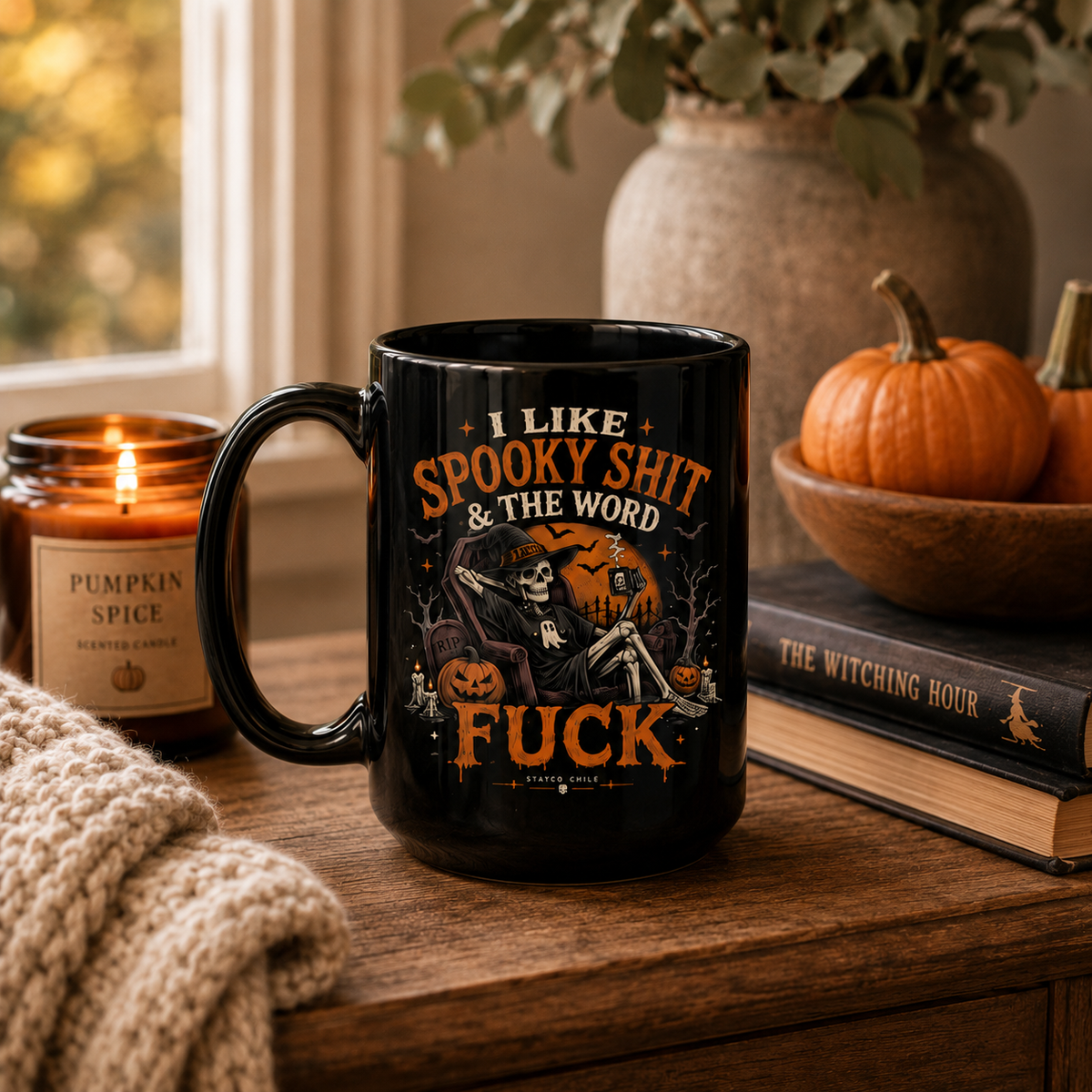 A black ceramic mug with a Halloween-themed graphic and text sits on a wooden table in a cozy indoor setting. The mug features an illustration of a skeleton under a full moon with pumpkins and the phrase 'I LIKE SPOOKY SHIT & THE WORD FUCK'.