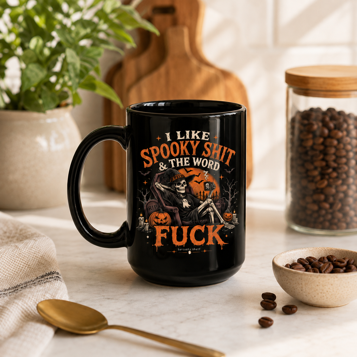 A black ceramic coffee mug with a Halloween-themed graphic print, styled on a marble countertop with coffee beans, a plant, and kitchen props in the background.