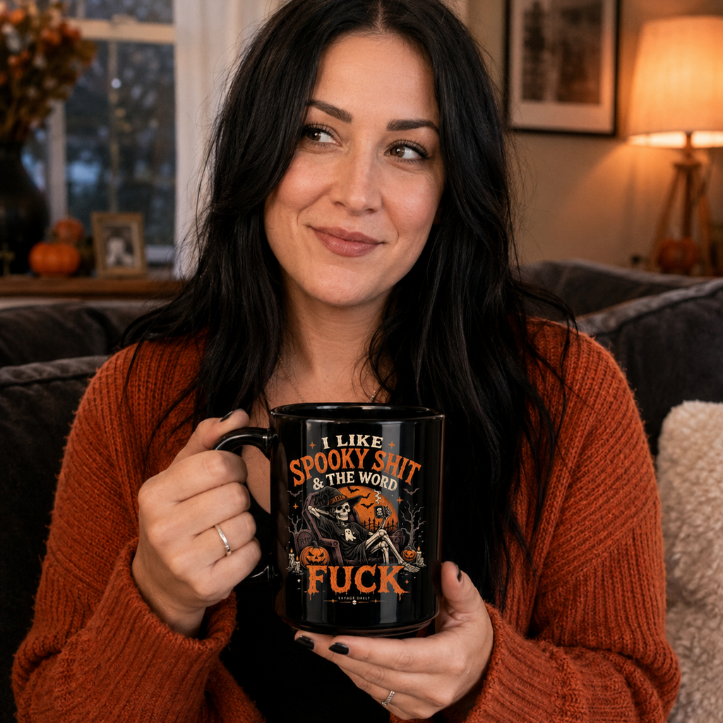 A woman with long dark hair sits on a couch holding a black ceramic mug with a Halloween-themed graphic and the text 'I LIKE SPOOKY SHIT THE WORLD FUCK'. She is wearing a rust-orange knit cardigan sweater in a cozy living room setting.